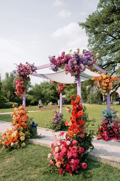 Wedding ceremony arch with colorful floral wedding arch roses, purple blooms, and greenery, draped fabric on a garden lawn by trees and a house exterior