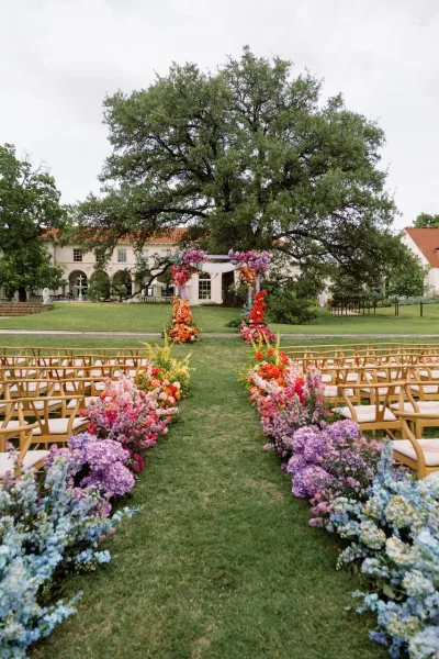 Ceremony setup for an outdoor wedding ceremony with a floral arch and fabric draping, flower-lined aisle, wooden chairs on a lawn by an estate under cloudy sky