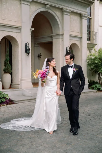 Couple portrait of bride and groom walking hand in hand, bride with bright bouquet and long veil by a stone archway courtyard