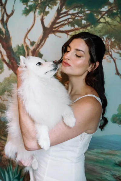 Bride with dog cuddling a fluffy white dog in a spaghetti strap wedding dress, posed before a painted landscape backdrop with trees