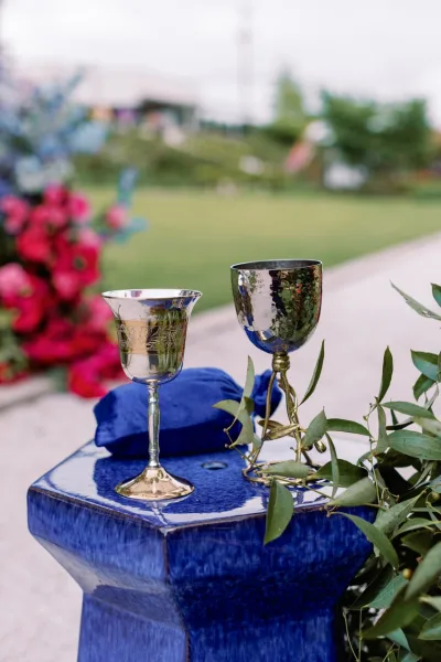 Wedding goblets and silver wedding goblets beside a blue velvet ring box on a blue pedestal with greenery garland and pink flowers outdoors