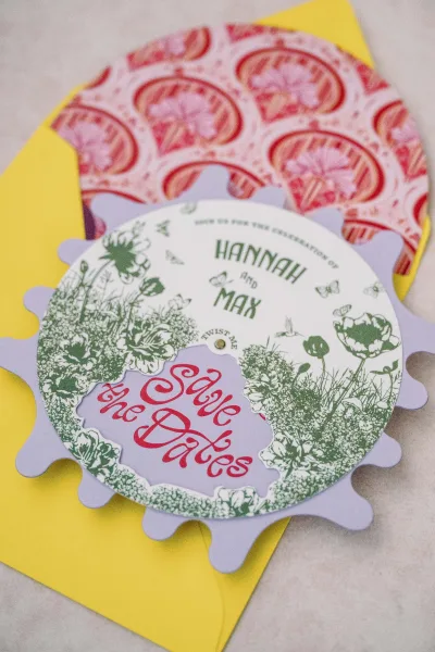 Save the date card with die-cut circular scalloped edge and floral illustration, layered with envelope and inserts on stone surface