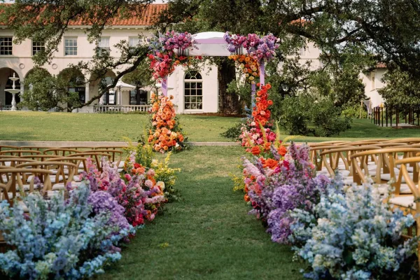 Ceremony altar decor with a colorful floral ceremony arch, pastel aisle flowers and ribbon on a lawn before a white estate building