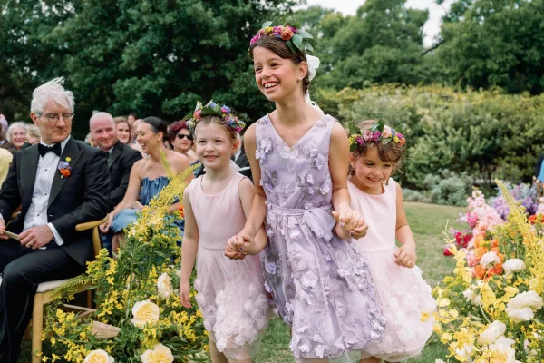 Flower girls holding hands in blush and lavender dresses with floral crowns, walking down a garden aisle past seated guests