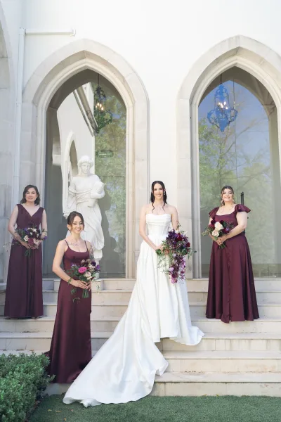 Bride with bridesmaids portrait on stone steps, bride in strapless gown holding cascading bouquet beside bridesmaids in burgundy dresses