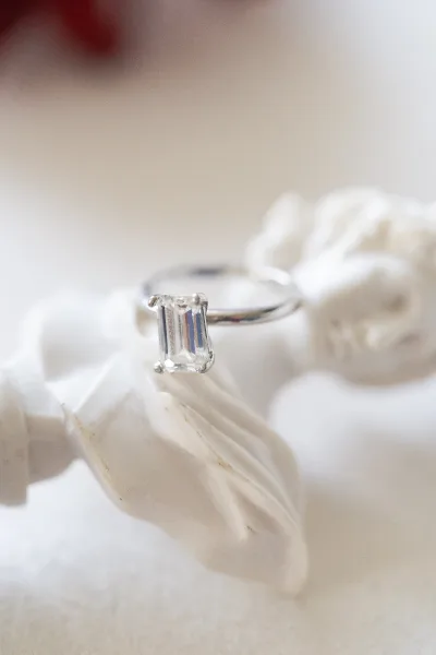Engagement ring with an emerald cut diamond rests on white flower petals over a soft neutral surface with blurred floral background