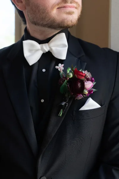 Groom portrait in a black tuxedo groom look with white bow tie, boutonniere, and satin lapels lit by soft window light indoors