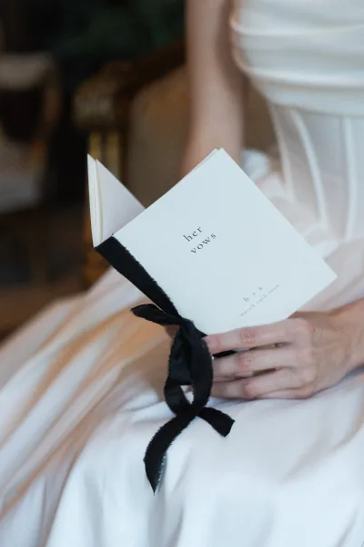 Wedding vows book with handwritten wedding vows tied in a black ribbon, held in the bride’s hands against a warm indoor background