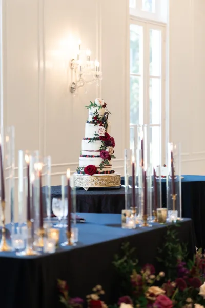Wedding cake on a gold cake stand, tiered with fresh flowers and berries, flanked by tall taper candles on black linens in a bright room