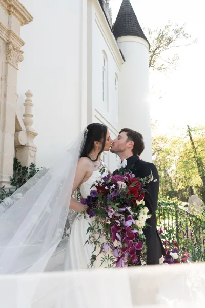 Wedding kiss as bride in strapless dress and veil holds bouquet, groom in tuxedo by a sunlit castle-like exterior with turret