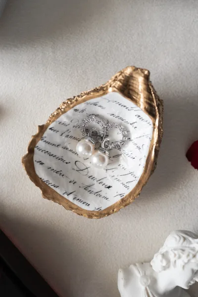 Bridal earrings with pearl drop earrings in a gold shell dish beside calligraphy paper on linen, styled with a red petal and figurine