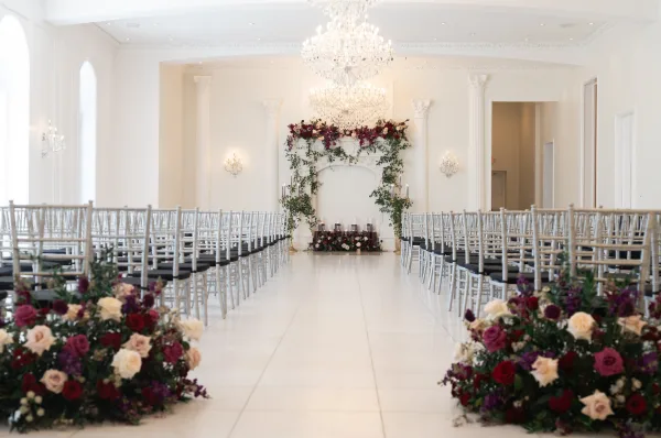 Ceremony setup with an indoor wedding ceremony aisle, white runner lined with florals and candles leading to a burgundy-blush arch in a bright white room