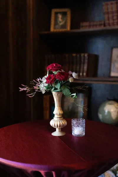 Wedding centerpiece with a red rose centerpiece in a gold vase and votive candle, set on a burgundy tablecloth before library bookshelves