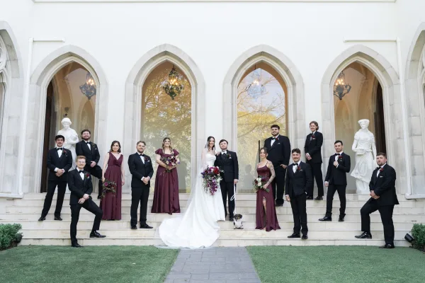 Wedding party portraits with bride and groom lined up on stone steps, bride in long train and veil, bridesmaids in burgundy by arched windows