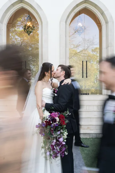 Wedding kiss portrait of bride and groom kissing in an embrace, her veil trailing and purple rose bouquet before arched windows and chandelier reflection