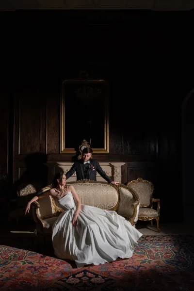 Couple portrait of bride on vintage sofa in a strapless wedding dress, groom in black tuxedo behind her in a dark wood room