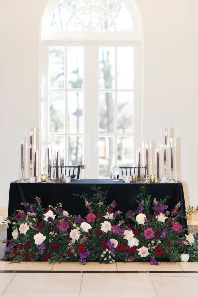 Sweetheart table decor with black tablecloth, roses and greenery ground arrangement, tall taper candles and gold candlesticks by an arched window