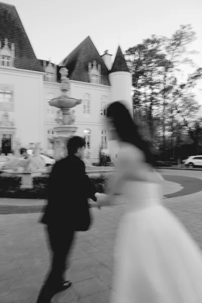 Couple portrait in a black and white wedding portrait, bride and groom holding hands as they walk away past a courtyard fountain and castle building