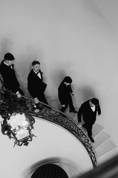 Groomsmen portrait of men in black tuxedos and bow ties walking upstairs on a curved staircase with wrought iron railing and chandelier above
