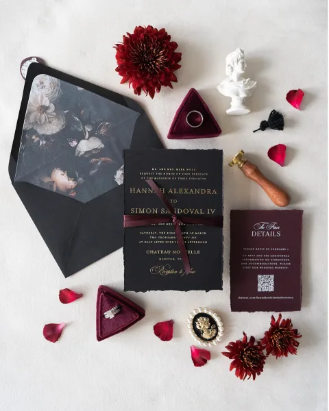 Invitation suite with black wedding invitations, floral-lined envelope, velvet ring box and rings, burgundy ribbon and wax seal on white fabric backdrop