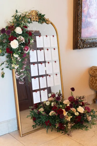 Wedding seating chart on a gold mirror with calligraphy escort cards and wax seals, framed by roses and greenery against a white wall