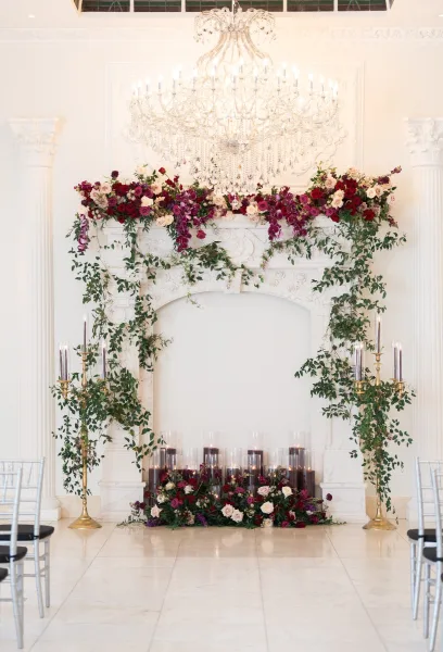 Wedding ceremony backdrop with fireplace wedding backdrop, roses and greenery garland on a white mantel beneath a crystal chandelier and candles