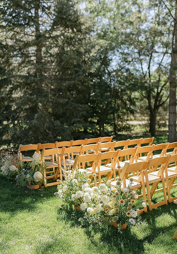 Ceremony seating with outdoor wedding chairs lined on a grass aisle, accented by white-and-green ground florals in a tree-filled garden setting