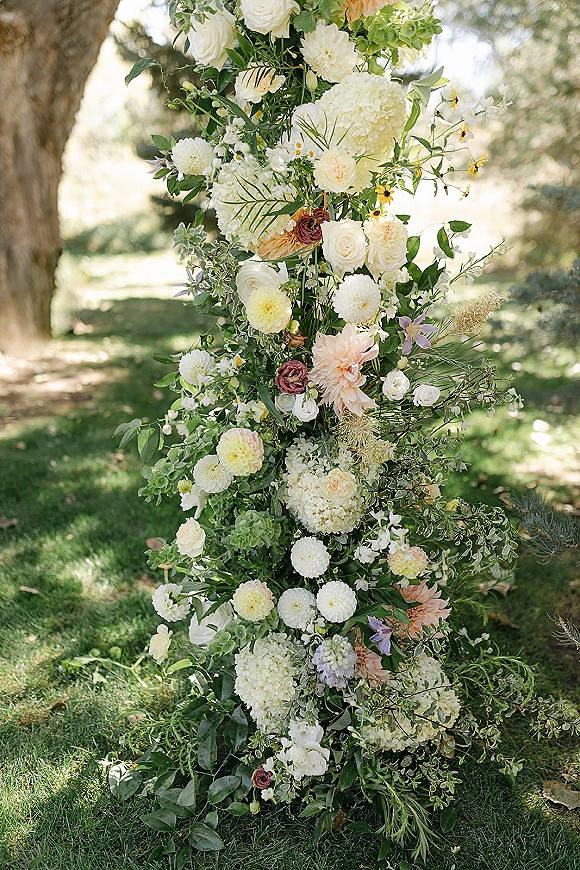 Wedding ceremony flowers on a floral arch arrangement with roses, dahlias, and hydrangea, framed by greenery in a sunlit garden lawn