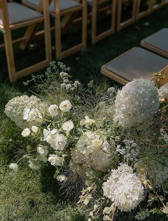 Ceremony aisle flowers with white hydrangeas and garden roses in airy greenery, arranged as a floral meadow runner beside wood chairs on grass lawn