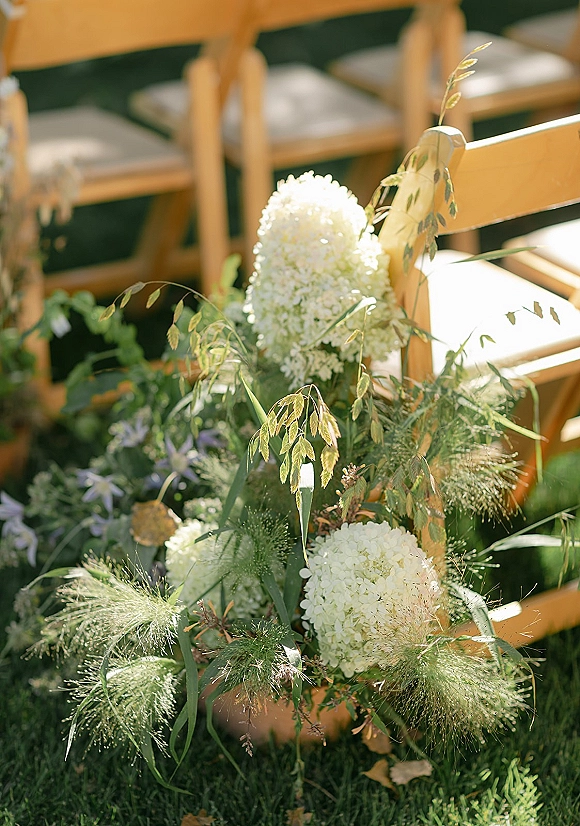 Ceremony aisle flowers with white hydrangeas and greenery nestled in ornamental grasses beside wooden chairs on a sunlit lawn