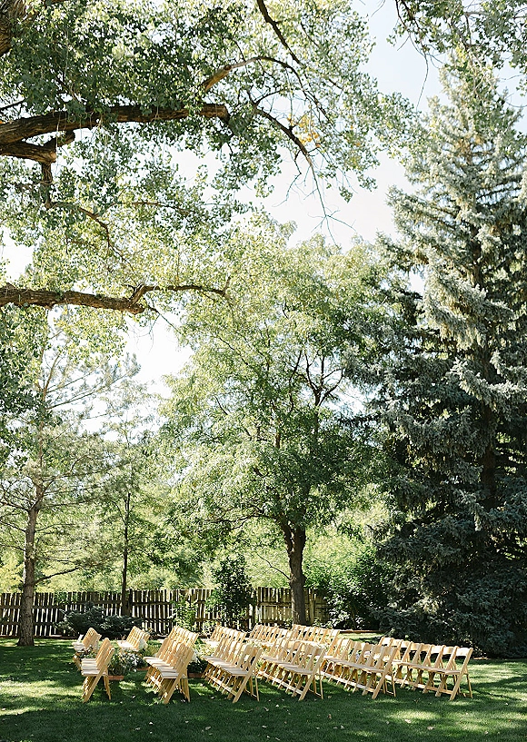Outdoor ceremony setup with backyard wedding ceremony chair rows of wood folding chairs and aisle florals on a sunny lawn under trees