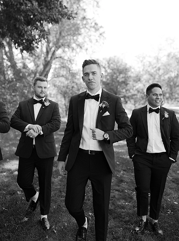 Groomsmen portrait of men in black tuxedos with bow ties and boutonnieres standing together on a grassy park lawn under trees