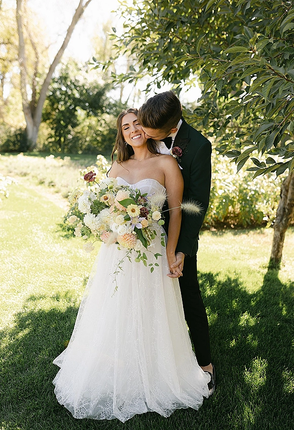 Couple portrait of bride and groom kissing, holding hands as he kisses her cheek on a sunlit garden lawn beneath leafy trees