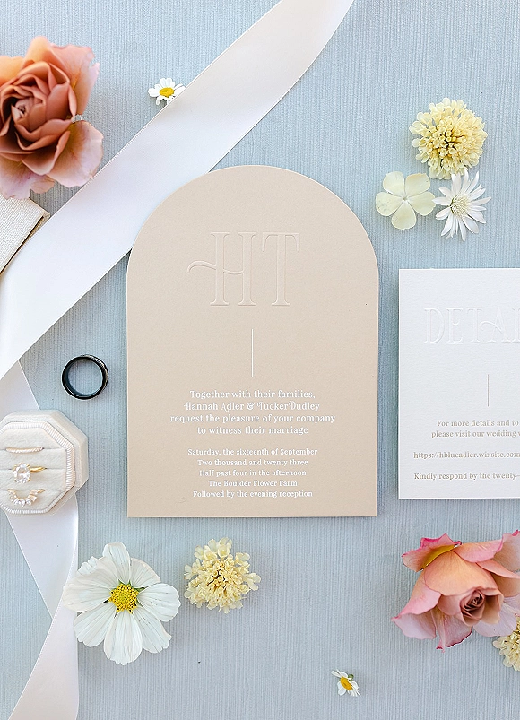Invitation suite with an arched wedding invitation, white ribbon, ring box, bands, and roses on a light blue textured surface