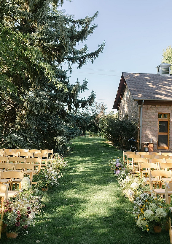 Ceremony aisle decor with bucket flower arrangements and greenery lining wood folding chairs on a lawn beside a stone building under blue sky