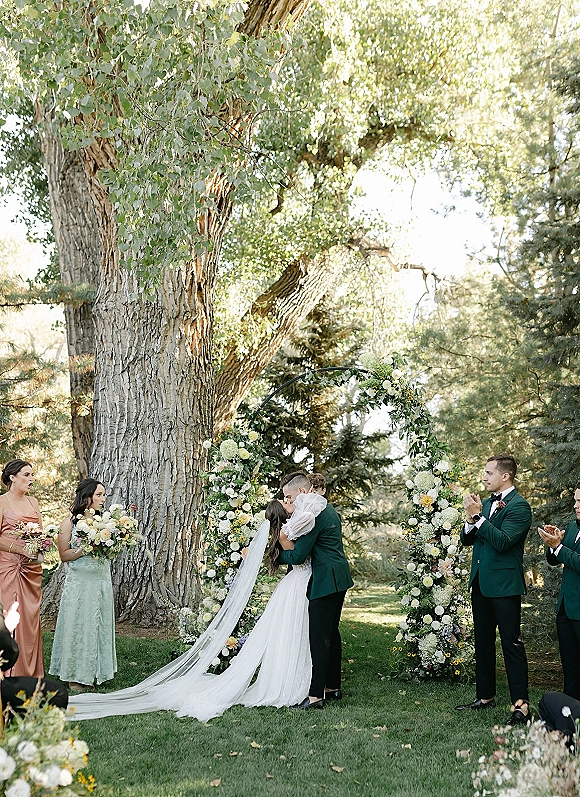 Wedding kiss beneath a white-and-green floral arch as the bride’s cathedral veil trails on a garden lawn while groomsmen clap