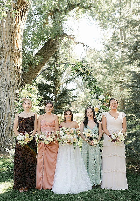 Bridesmaid group photo with bride and bridesmaids holding bouquets beneath a circular floral arch on a sunlit park lawn
