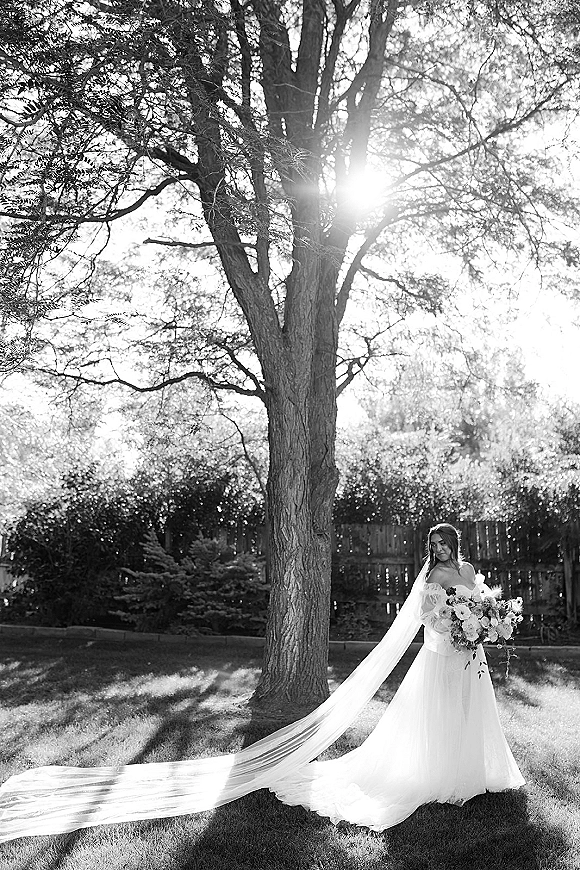 Bridal portrait of a bride holding bouquet with a long veil, standing on a garden lawn under a tree with sun flare and fence behind