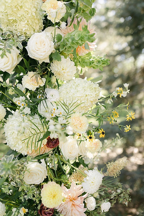Wedding floral arrangement with white roses and hydrangea, accented by yellow wildflowers and greenery against sunlit trees bokeh