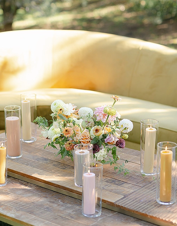 Reception centerpiece with low wedding centerpiece of pastel blooms, greenery, and pillar candles in glass cylinders on a wooden lounge coffee table outdoors