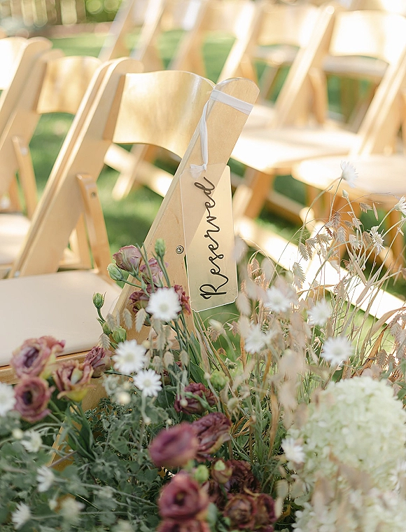 Ceremony seating decor with wood folding chairs, a calligraphy chair tag and ribbon, plus daisies and hydrangea on a grass lawn outdoors