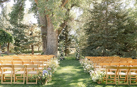 Outdoor ceremony setup with wood folding chairs lining a wedding aisle, leading to a floral arch on a green lawn beneath tall trees