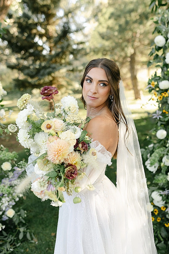 Bridal portrait of a bride holding bouquet, looking over her shoulder in an off-the-shoulder lace gown with veil on a garden lawn