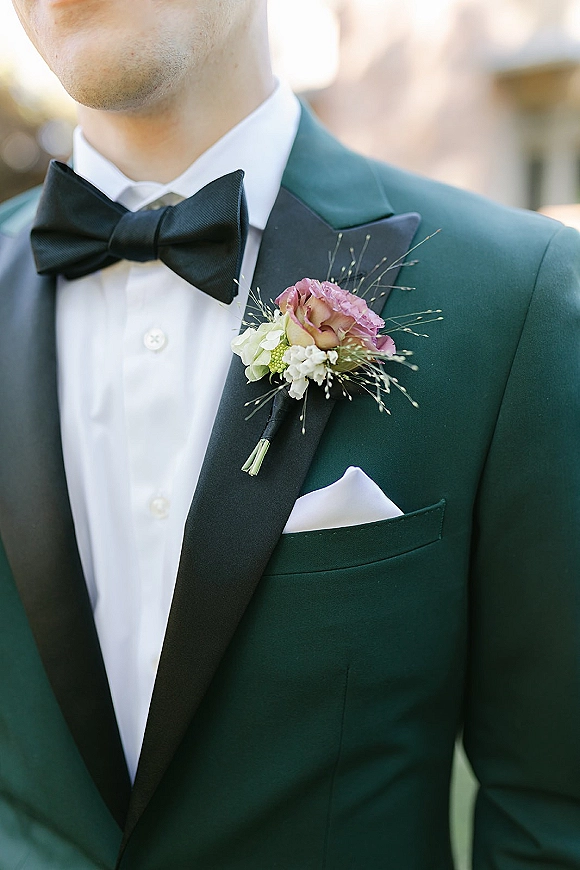 Groom boutonniere with a rose boutonniere and greenery pinned to a tuxedo lapel, paired with a black bow tie and white pocket square outdoors