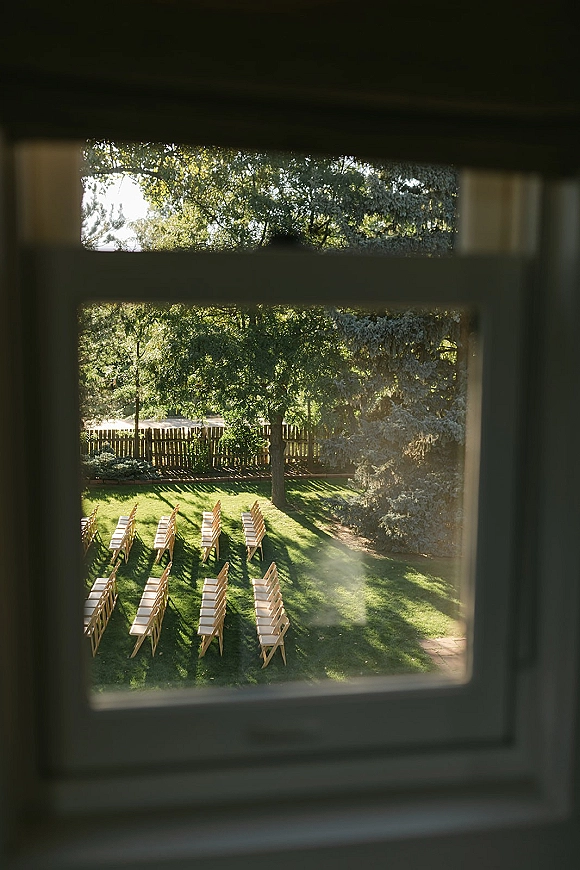 Ceremony seating with wood folding chairs arranged in neat rows on a grass lawn, framed by trees, a wooden fence, and a window view