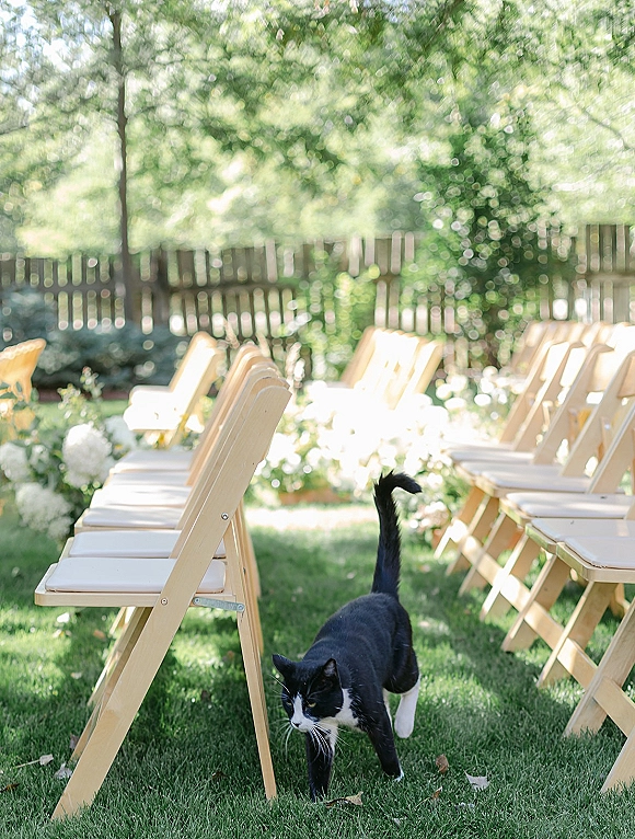 Ceremony seating with wood folding chairs and white cushions arranged in rows on a grass lawn, lined with white floral accents by the aisle