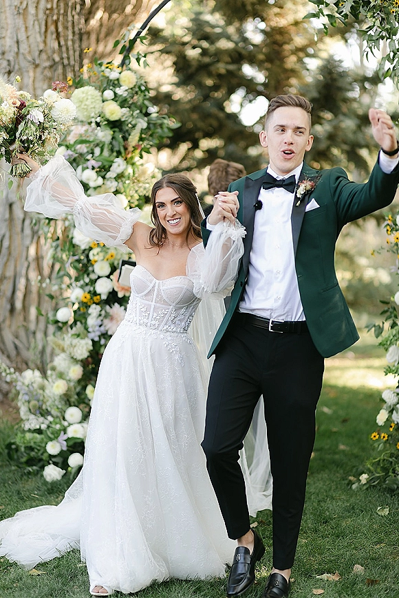 Wedding recessional as bride and groom walking aisle, holding hands under a floral arch, bride lifts bouquet overhead on garden lawn