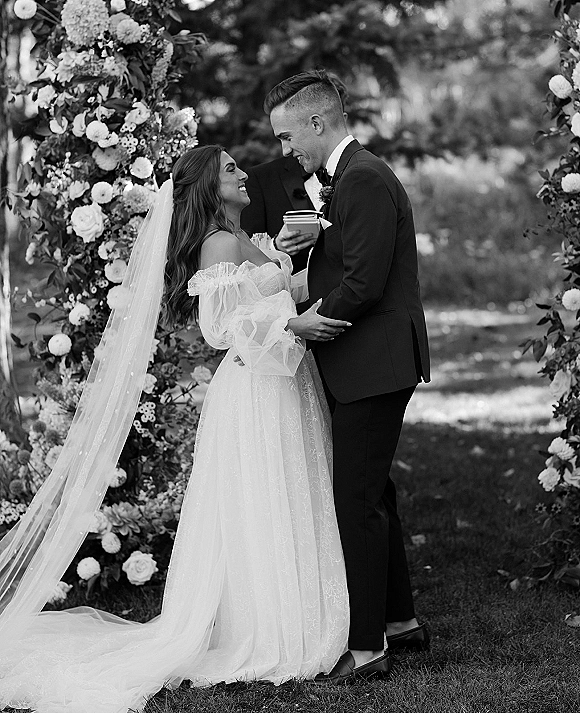 Wedding ceremony moment as bride and groom smile exchanging vows under a floral arch, veil trailing on a garden lawn with trees behind