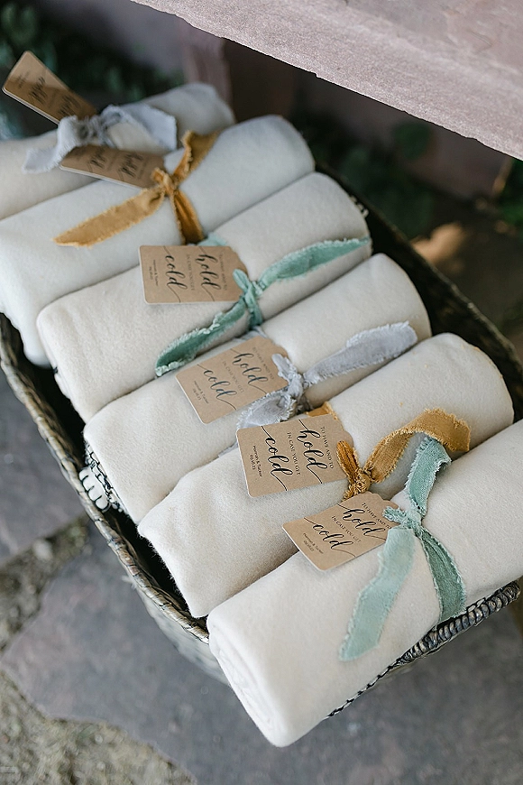 Wedding favors with rolled napkin favors tied in dusty blue and mustard ribbon, labeled name tags, arranged in a woven basket on stone floor with greenery