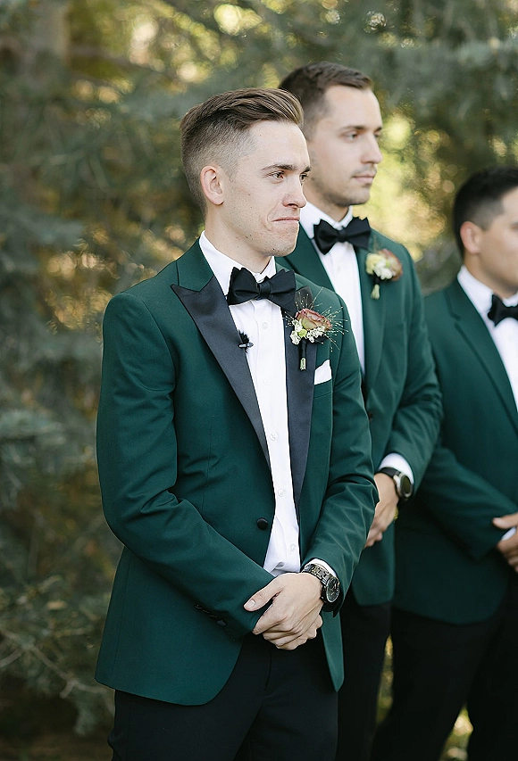 Groom portrait in an emerald green tuxedo, hands clasped with black bow tie, boutonniere, and pocket square against trees backdrop